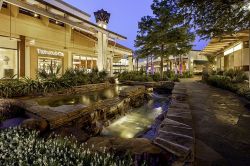 Outdoor mall with lit storefronts, stone pathway, and small cascading water feature surrounded by plants at dusk near Enclave at the Dominion.