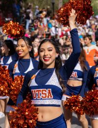 Cheerleaders in UTSA uniforms wave orange pom-poms, marching in a crowded parade near Enclave at the Dominion.