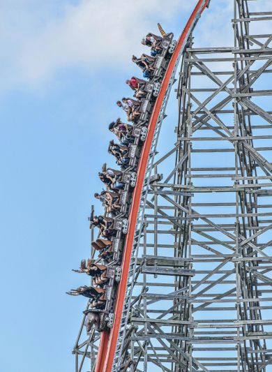 People riding a steep roller coaster drop on a wooden structure against a blue sky near Enclave at the Dominion apartments.