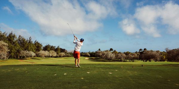 Enclave at the Dominion Golfer in red shorts and white shirt swings on a lush green course under a partly cloudy sky.