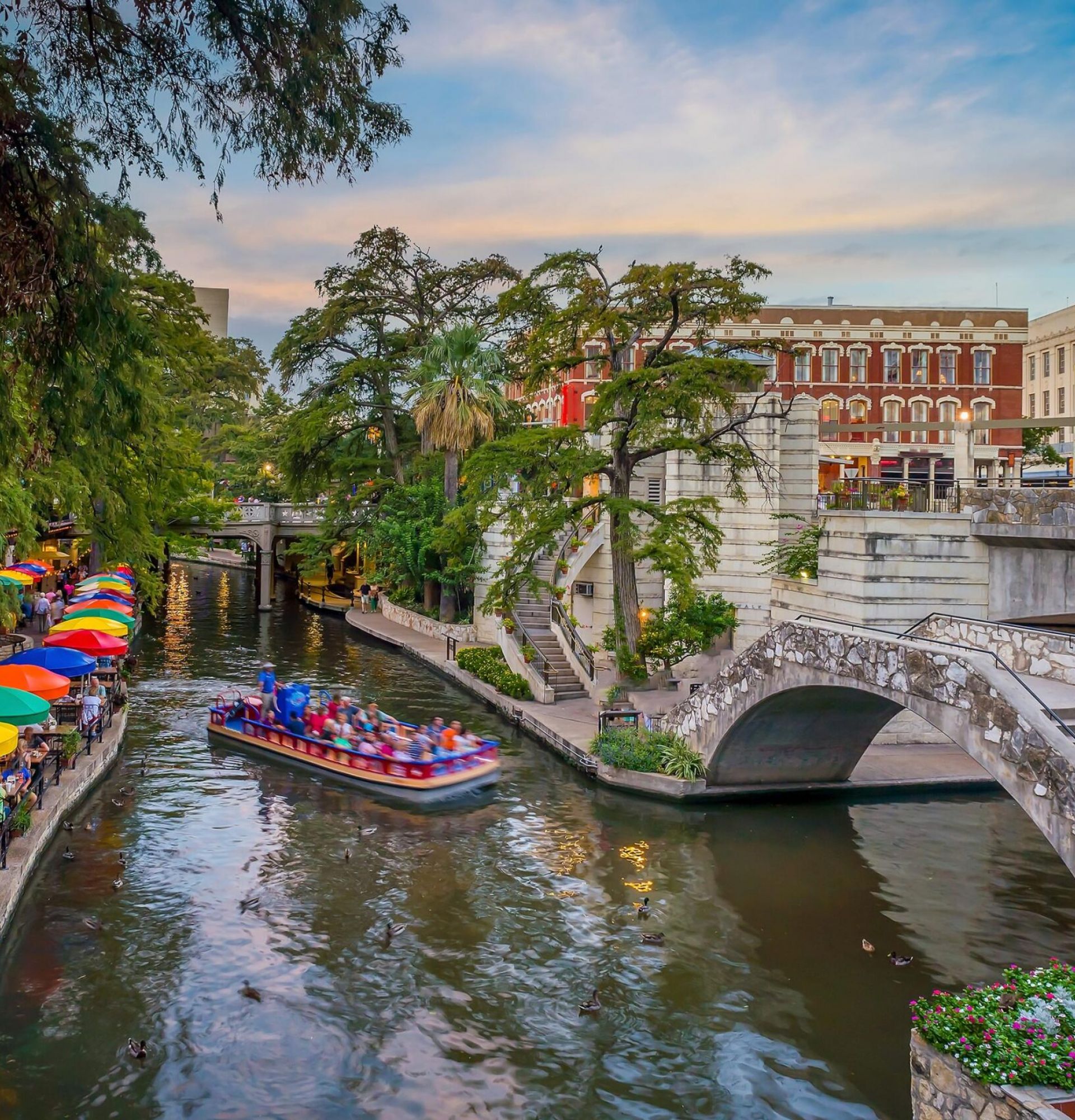 Enclave at the Dominion Colorful umbrellas line the San Antonio River Walk as a boat passes under a stone bridge.