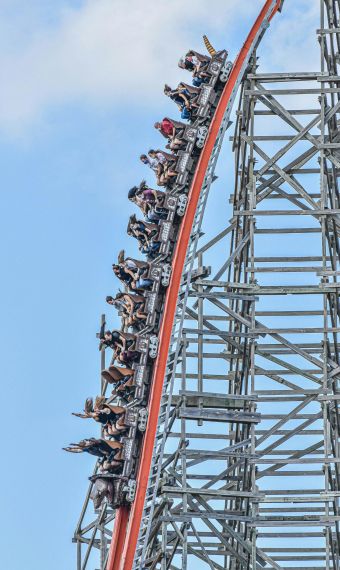 People riding a steep roller coaster drop on a wooden structure against a blue sky near Enclave at the Dominion apartments.