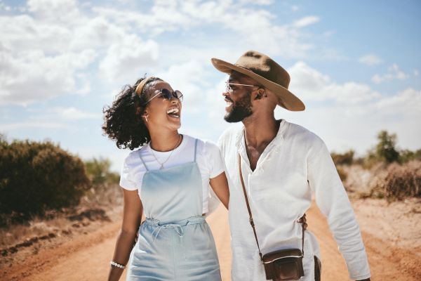 A smiling couple walks arm-in-arm down a sunny dirt road, both wearing sunglasses and casual summer clothes near Enclave at the Dominion apartments.